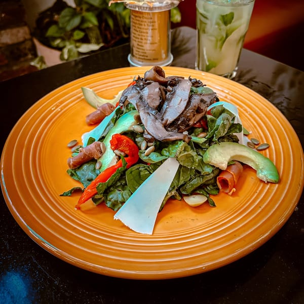 A vibrant Spanish salad with mushrooms, avocado, prosciutto, and cheese on an orange plate.